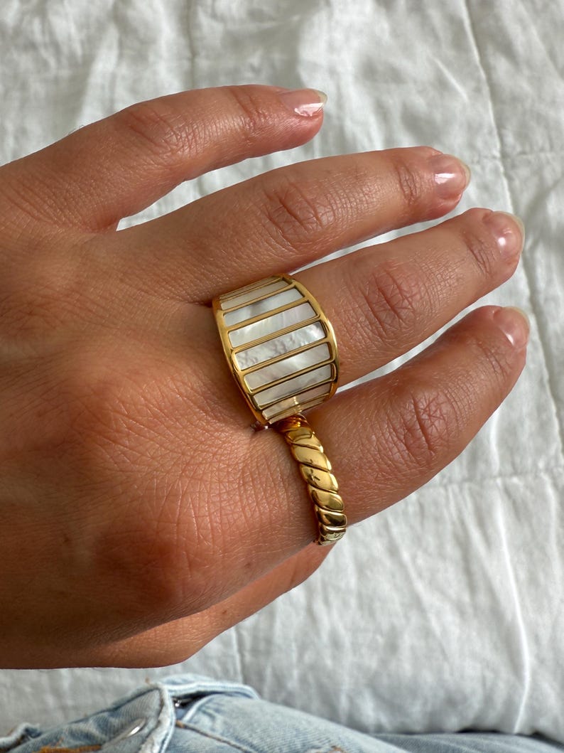 May include: A wide gold-tone ring featuring vertical inlays of iridescent white mother-of-pearl segments. The band is connected to a secondary, thinner gold-tone ring with a twisted, rope-like texture that sits lower on the finger.