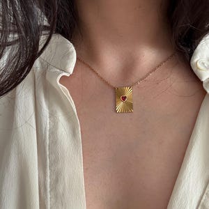 Pode incluir: Colar dourado com pingente retangular e coração vermelho. O pingente tem um design de raios de sol. O colar é usado sobre uma camisa branca.