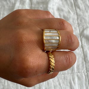 May include: A wide gold-toned ring featuring vertical panels of iridescent white mother-of-pearl inlay, paired with a second gold-toned ring that has a twisted, rope-like texture. Both rings are worn on the fingers, showcasing their metallic finish.