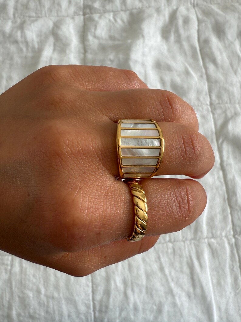 May include: A wide gold-toned ring featuring vertical panels of iridescent white mother-of-pearl inlay, paired with a second gold-toned ring that has a twisted, rope-like texture. Both rings are worn on the fingers, showcasing their metallic finish.