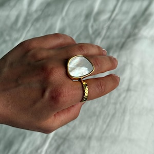May include: A gold-toned ring featuring a large, irregular, pearlescent white stone set in a smooth bezel. The band of the ring has a distinct twisted or rope-like texture, providing a contrast to the polished, iridescent surface of the central stone.