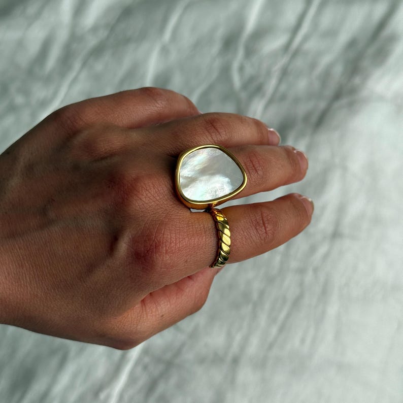Puede incluir: Un anillo de tono dorado con una piedra blanca nacarada grande e irregular engastada en un bisel liso. La banda del anillo tiene una textura trenzada o similar a una cuerda, lo que contrasta con la superficie pulida e iridiscente de la piedra.