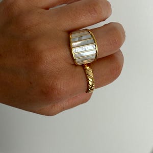 May include: A gold-toned ring featuring a wide, curved band with vertical inlays of iridescent white mother-of-pearl, paired with a second gold-toned ring that has a twisted, rope-like texture on the band.