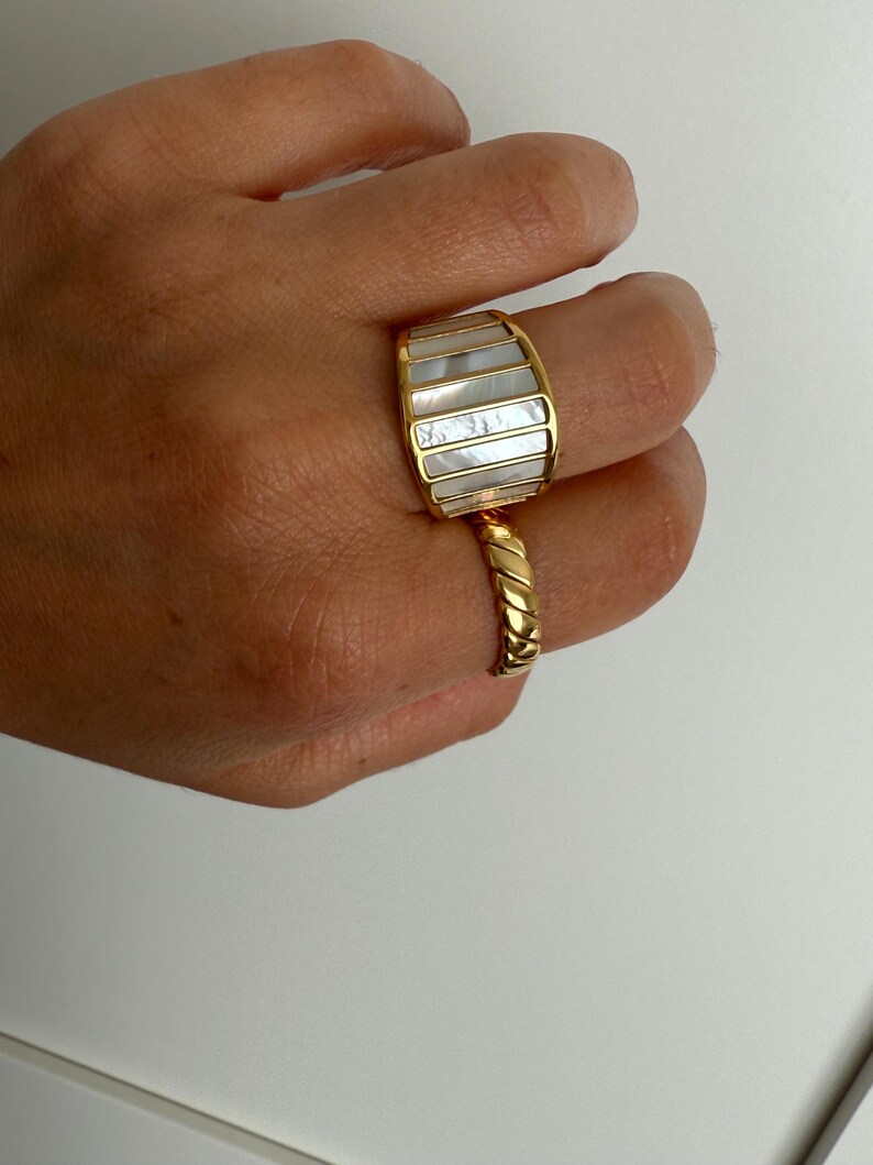 May include: A gold-toned ring featuring a wide, curved band with vertical inlays of iridescent white mother-of-pearl, paired with a second gold-toned ring that has a twisted, rope-like texture on the band.