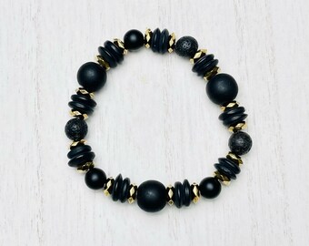 May include: Black beaded bracelet with gold accents. The bracelet features round black beads, flat black beads with gold trim, and small black lava rock beads.