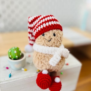 May include: A tan crochet plush figure with a textured surface, wearing a red and white striped hat with a white pom-pom, a white fuzzy scarf, and red crochet shoes with white pom-poms. The figure has a stitched face with black eyes.