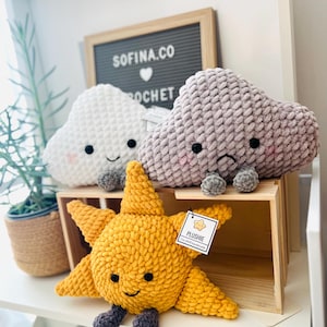 May include: A display of handmade crochet plushies, including a yellow star, a white cloud, and a gray cloud. The star has a smiling face and gray feet. The clouds have smiling and frowning faces, with gray feet. The sign says "SOFINA.CO CROCHET".