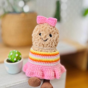 May include: A tan plush doll with a fuzzy texture, featuring a pink crocheted bow on its head and a silver beaded necklace. The doll wears a dress with horizontal stripes in yellow, orange, and white, finished with a ruffled pink skirt and brown feet.
