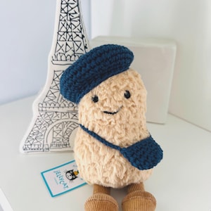 May include: A plush toy with a fuzzy beige body, wearing a dark blue crocheted beret and a matching dark blue crocheted crossbody bag. The toy has small black eyes, a stitched smile, and brown corduroy legs. A Jellycat brand tag is attached.