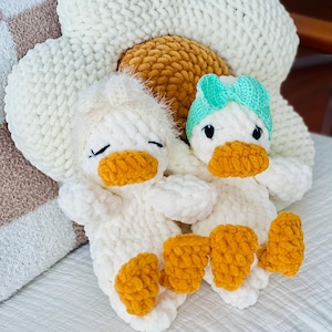 May include: Two white crochet duck plushies with orange beaks and feet, one wearing a teal bow. They are resting on a large white crochet flower pillow with a yellow center. The ducks are on a white surface.