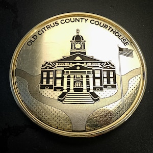 Puede incluir: Una moneda plateada con el antiguo juzgado del condado de Citrus. La moneda tiene un grabado detallado del edificio del juzgado, una bandera estadounidense y el texto "OLD CITRUS COUNTY COURTHOUSE" alrededor del borde.