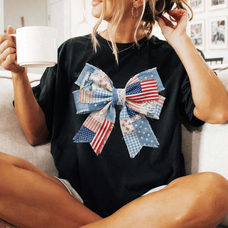 May include: Black t-shirt with a large bow design. The bow is made from denim, lace, and patches with American flag and star patterns. The shirt is a casual style.