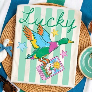 May include: Cream-colored t-shirt with a colorful duck graphic holding a tarot card that reads "The Sun." The word "Lucky" is written in green script above the duck. The shirt has vertical mint green stripes and star accents.