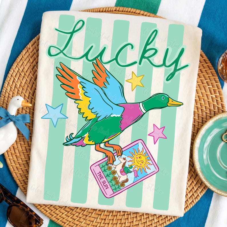 May include: Cream-colored t-shirt with a colorful duck graphic holding a tarot card that reads "The Sun." The word "Lucky" is written in green script above the duck. The shirt has vertical mint green stripes and star accents.