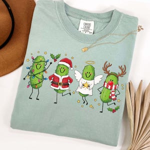 May include: A sage green t-shirt featuring a graphic of four cartoon pickles dressed for the holidays. One is wrapped in colourful string lights, one wears a Santa suit, one is an angel with wings and a halo, and one has reindeer antlers.