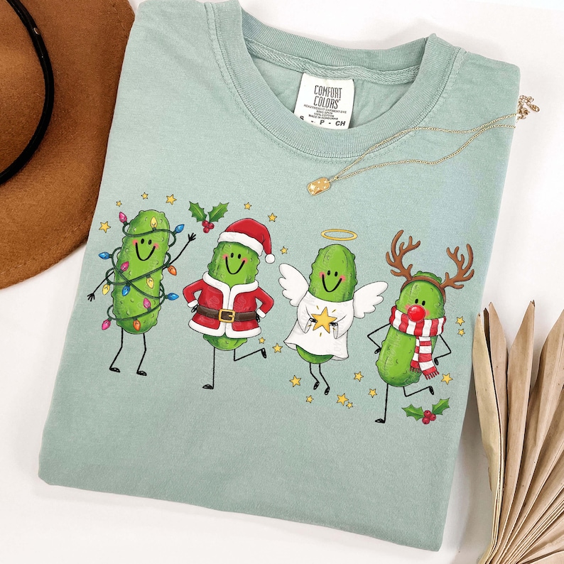May include: A sage green t-shirt featuring a graphic of four cartoon pickles dressed for the holidays. One is wrapped in colourful string lights, one wears a Santa suit, one is an angel with wings and a halo, and one has reindeer antlers.