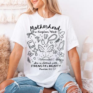 May include: White t-shirt with a black graphic design that says "Motherhood is Kingdom Work" and includes various religious symbols like a cross, dove, bible, and angel wings. The text "She is clothed with STRENGTH & DIGNITY Proverbs 31:25" is also included in the design.