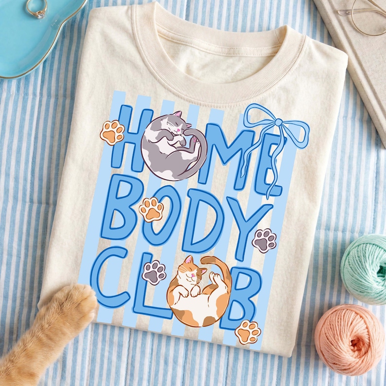 May include: Cream-colored t-shirt with the text "HOME BODY CLUB" in blue, featuring illustrations of two cats and paw prints. The shirt is on a blue and white striped surface, with yarn balls and a ring in the background.
