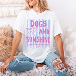 May include: White t-shirt with the text "DOGS AND SUNSHINE" in pink, with a paw print and bow graphic. The shirt also includes the phrase "enjoying the simple things" in a script font. The shirt is paired with light blue ripped jeans.