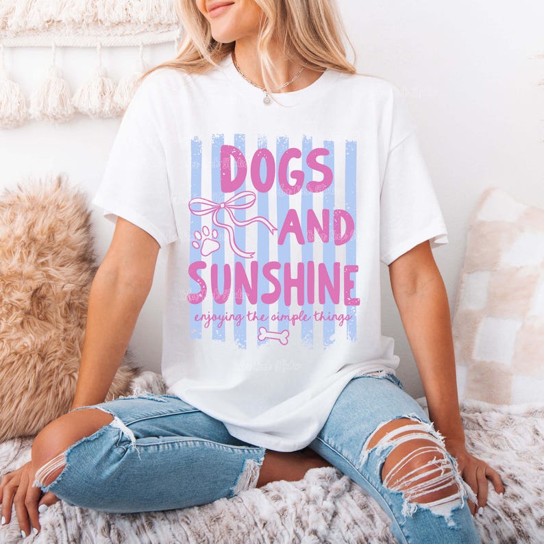 May include: White t-shirt with the text "DOGS AND SUNSHINE" in pink, with a paw print and bow graphic. The shirt also includes the phrase "enjoying the simple things" in a script font. The shirt is paired with light blue ripped jeans.