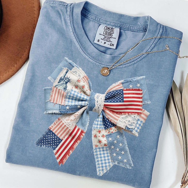 May include: A pale blue Comfort Colors t-shirt featuring a large bow made from fabric patches. The bow design includes American flag patterns, floral prints, and lace. A gold necklace with a pendant is draped on the shirt.