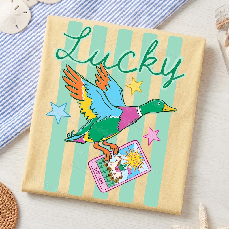 May include: A tan-colored towel with vertical light green stripes features a colorful cartoon duck with the word "Lucky" in green script. The duck holds a tarot card that says "The Sun".