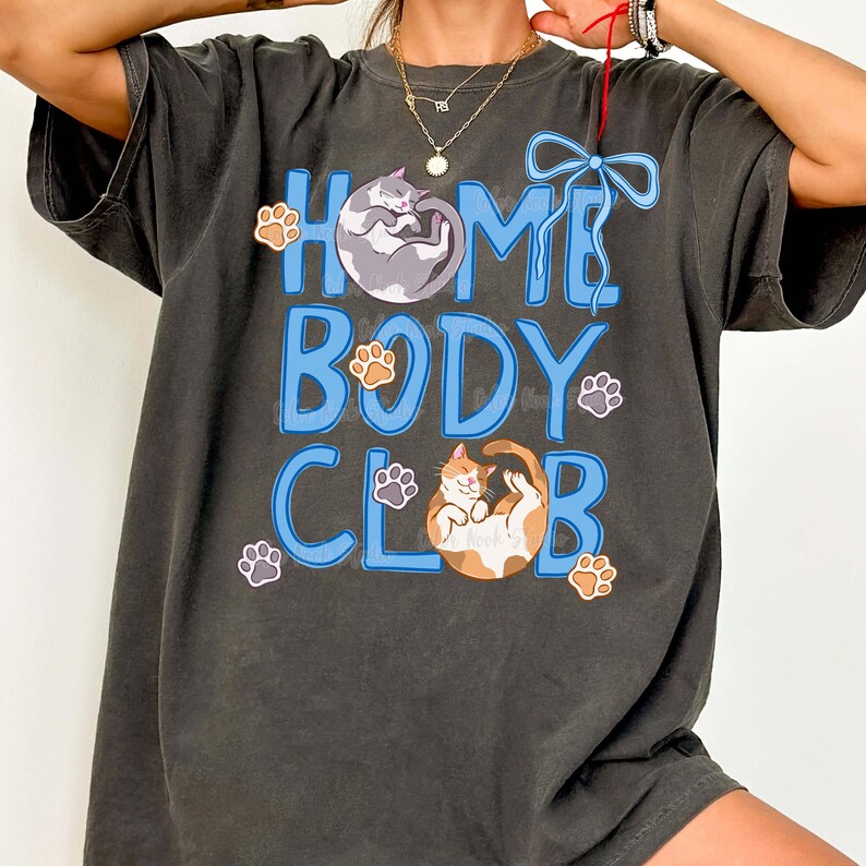 May include: Dark gray t-shirt with the words "HOME BODY CLUB" in blue, featuring illustrations of cats and paw prints. The shirt has a relaxed fit and a vintage look.