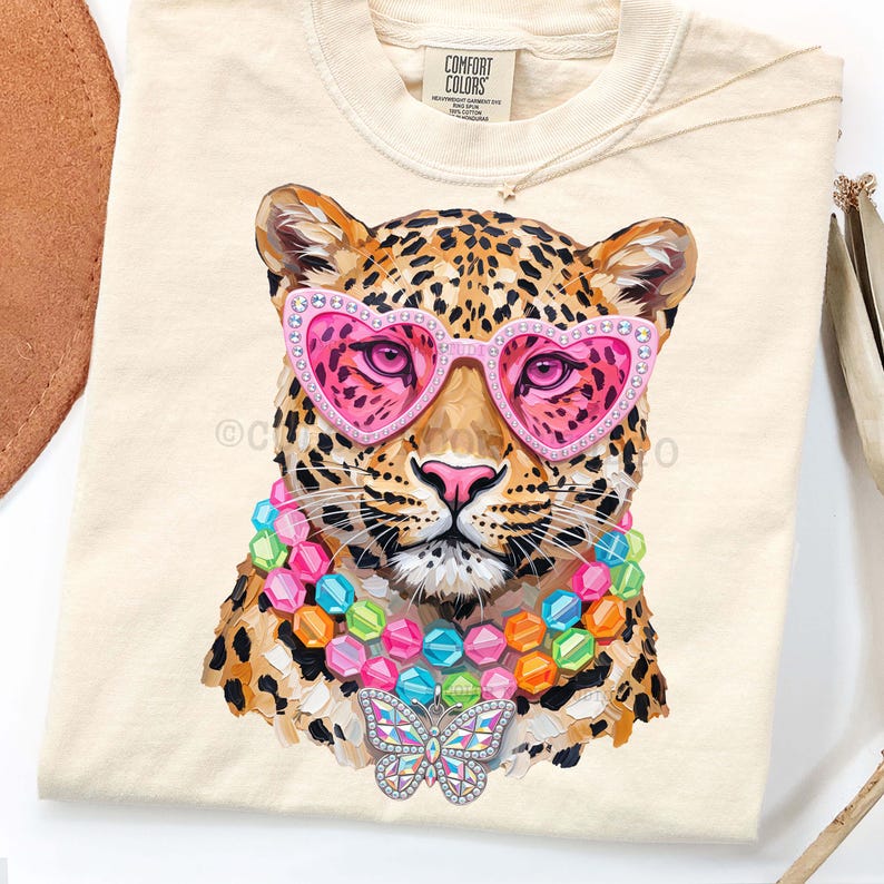 May include: A cream-colored t-shirt featuring a graphic of a leopard wearing pink heart-shaped sunglasses, a multi-colored beaded necklace, and a butterfly pendant. The shirt has a Comfort Colors brand label at the inner neckline.