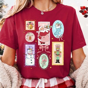 May include: Burgundy-colored t-shirt featuring a collage of Christmas-themed illustrations. The designs include a bell, Santa Claus, a snowman, a stocking, a gingerbread man, a candy cane, and a nutcracker. Each image is framed within a decorative border.