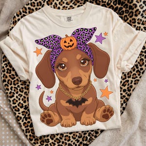 Puede incluir: Una camiseta color crema con el gráfico de un cachorro de perro salchicha marrón que lleva una diadema de estampado de leopardo morado con una calabaza, un collar de cadena dorada con un colgante de murciélago negro, rodeado de estrellas naranjas y moradas.