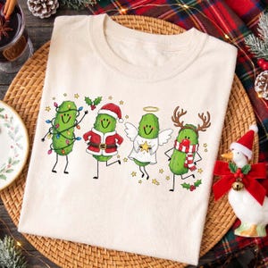 May include: A cream-coloured t-shirt featuring a graphic print of four green pickles dressed for the holidays. One pickle is wrapped in colourful Christmas lights, one wears a Santa suit, one is an angel with wings, and one has reindeer antlers.