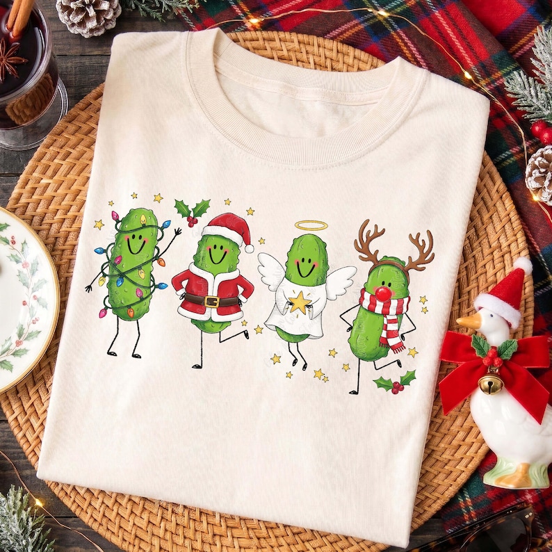 May include: A cream-coloured t-shirt featuring a graphic print of four green pickles dressed for the holidays. One pickle is wrapped in colourful Christmas lights, one wears a Santa suit, one is an angel with wings, and one has reindeer antlers.