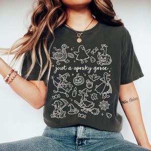May include: Dark gray t-shirt with a white graphic of Halloween-themed geese and the text "just a spooky goose." The design includes geese in costumes, pumpkins, bats, and witch hats. The shirt is a casual, unisex style.