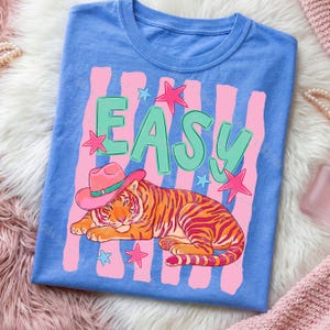 Puede incluir: Camiseta azul con un dise&ntilde;o gr&aacute;fico con la palabra "EASY" en letras verdes con estrellas rosas. Un tigre con un sombrero de vaquero rosa est&aacute; acostado. Rayas rosas y blancas en el fondo.