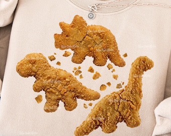May include: A beige sweatshirt featuring a trio of fried chicken dinosaur shapes. The dinosaurs are golden brown with a crispy texture, and small pieces of fried breading are scattered around them. A silver necklace is visible at the top.
