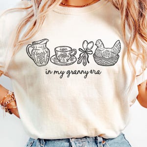 Puede incluir: Camiseta color crema con un dibujo lineal negro de una jarra, una taza de t&eacute;, cucharas y una gallina en una cesta. El texto "in my granny era" est&aacute; escrito en cursiva debajo. La camiseta tiene mangas cortas.