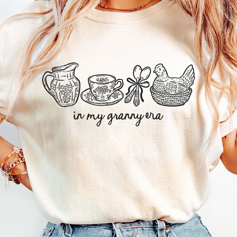 Puede incluir: Camiseta color crema con un dibujo lineal negro de una jarra, una taza de t&eacute;, cucharas y una gallina en una cesta. El texto "in my granny era" est&aacute; escrito en cursiva debajo. La camiseta tiene mangas cortas.