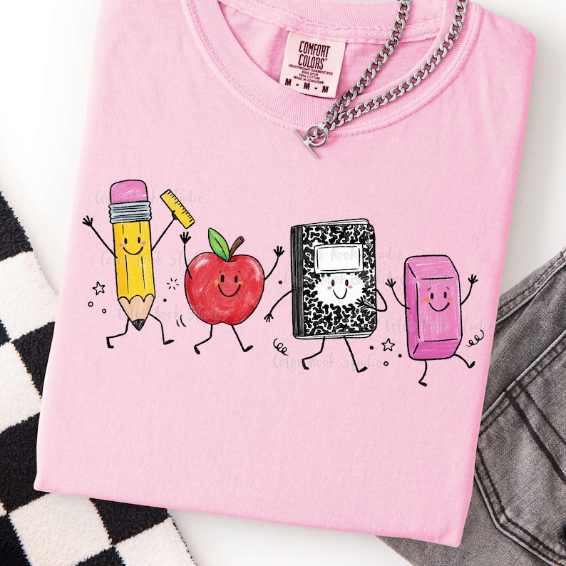 May include: Pink Comfort Colors t-shirt featuring a playful graphic of animated school supplies: a pencil, apple, notebook, and eraser. The shirt has a crew neck and short sleeves. A silver chain necklace is draped across the shirt.