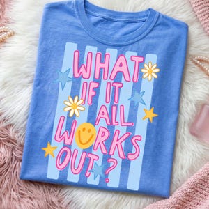 May include: A blue t-shirt with the phrase "WHAT IF IT ALL WORKS OUT?" in pink and white lettering. The design includes light blue stripes, stars, daisies, and a yellow smiley face. The shirt is folded on a fluffy surface.