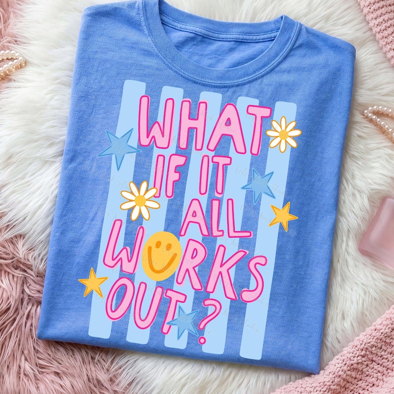 May include: A blue t-shirt with the phrase "WHAT IF IT ALL WORKS OUT?" in pink and white lettering. The design includes light blue stripes, stars, daisies, and a yellow smiley face. The shirt is folded on a fluffy surface.