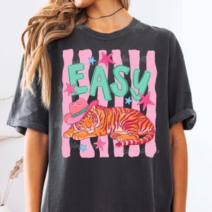 Puede incluir: Camiseta gris oscuro con un gr&aacute;fico de un tigre con un sombrero de vaquero rosa y la palabra "EASY" en verde azulado. Rayas verticales rosas y estrellas rodean al tigre. La camiseta es de estilo casual y vintage.