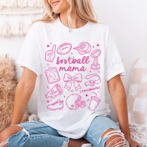 May include: White t-shirt with a pink graphic design that says "football mama" and includes various football-related icons, such as a football, a whistle, a cheerleader's bow, and a megaphone.