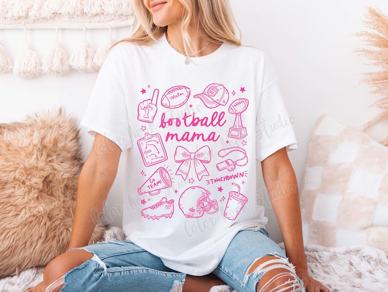 May include: White t-shirt with a pink graphic design that says "football mama" and includes various football-related icons, such as a football, a whistle, a cheerleader's bow, and a megaphone.