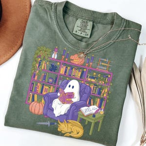 May include: Green Comfort Colors t-shirt with a graphic of a ghost reading a book. The design includes a bookshelf, pumpkin, and dragon. The text "Sorita Solis" is visible. A silver necklace is on the shirt.