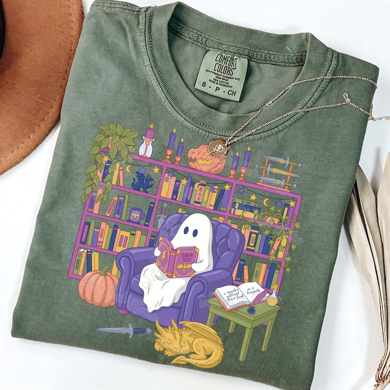 May include: Green Comfort Colors t-shirt with a graphic of a ghost reading a book. The design includes a bookshelf, pumpkin, and dragon. The text "Sorita Solis" is visible. A silver necklace is on the shirt.