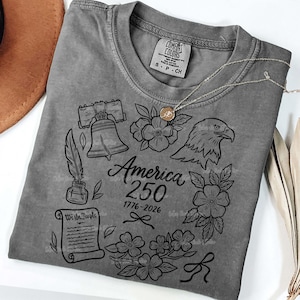 May include: A gray t-shirt with a graphic design commemorating America's 250th anniversary. The design includes the Liberty Bell, an eagle, flowers, a quill, and the text "America 250 1776-2026". A silver necklace with a pendant is draped over the shirt.