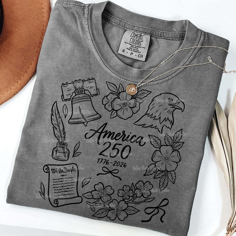 May include: A gray t-shirt with a graphic design commemorating America's 250th anniversary. The design includes the Liberty Bell, an eagle, flowers, a quill, and the text "America 250 1776-2026". A silver necklace with a pendant is draped over the shirt.