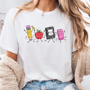 May include: White t-shirt featuring a playful graphic of animated school supplies: a pencil, apple, notebook, and eraser. The pencil is yellow with a pink eraser, the apple is red, the notebook is black and white, and the eraser is pink. The design has a cartoon style.
