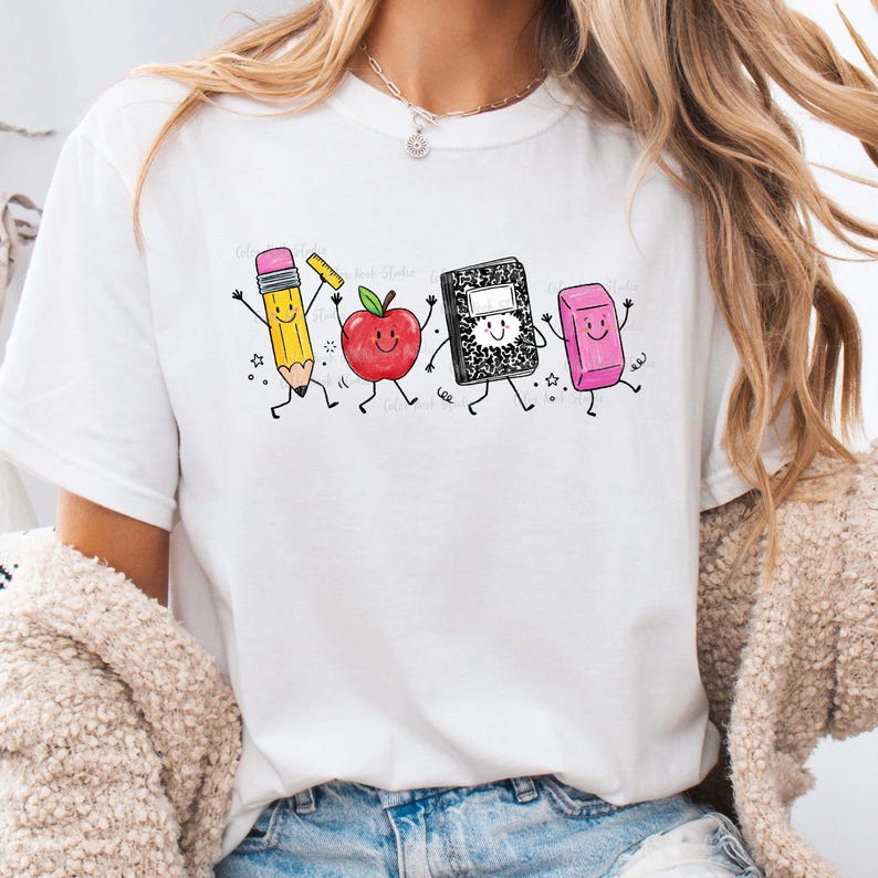 May include: White t-shirt featuring a playful graphic of animated school supplies: a pencil, apple, notebook, and eraser. The pencil is yellow with a pink eraser, the apple is red, the notebook is black and white, and the eraser is pink. The design has a cartoon style.