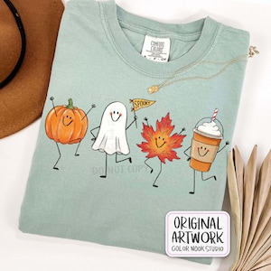 May include: A sage green t-shirt featuring a graphic of a dancing pumpkin, a ghost holding a yellow pennant with the text SPOOKY, a maple leaf, and a coffee cup with whipped cream. The shirt has a Comfort Colors label at the neck.